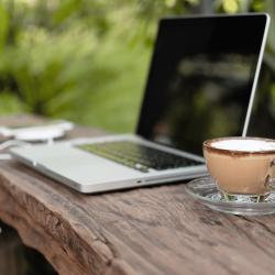 A laptop on a wooden desk outside with a cup of coffee